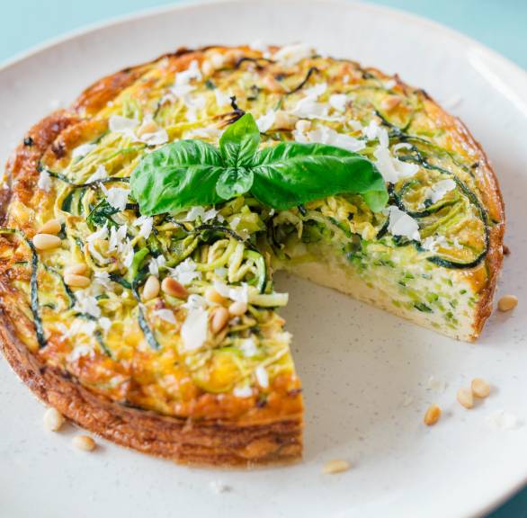 Runder Zucchini-Nudel-Kuchen angeschnitten auf einem Teller garniert mit Basilikum, daneben ein Stück Parmesan
