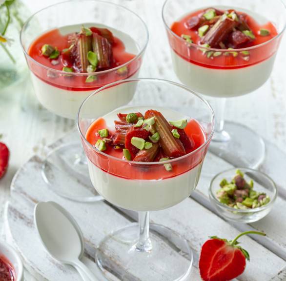 Erdbeer-Rhabarber-Panna Cotta in drei Gläsern getoppt mit Pistazien auf einem weißen Holztisch