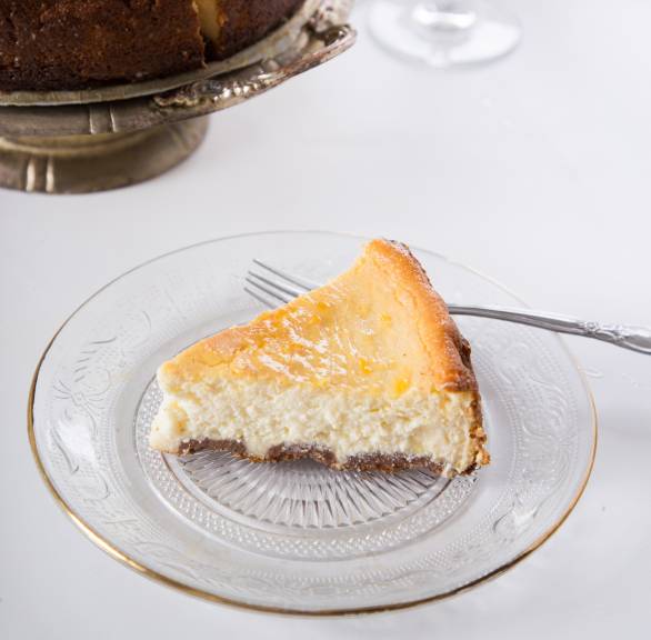 Ein Stück Ricotta-Cheesecake auf einem Kuchenteller, dahinter der restliche Kuchen auf einer Etagere