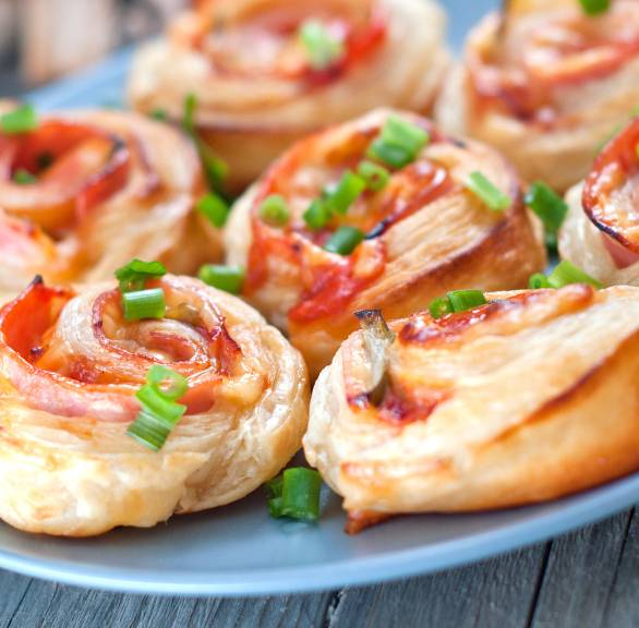 Sieben Pizzaschnecken aus Blätterteig auf einem Teller, Füllung aus Schinken und Käse sichtbar