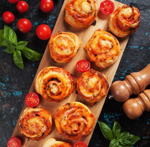 Neun Pizzaschnecken auf einem rechteckigen Holzbrett, daneben Tomaten auf einem Tisch