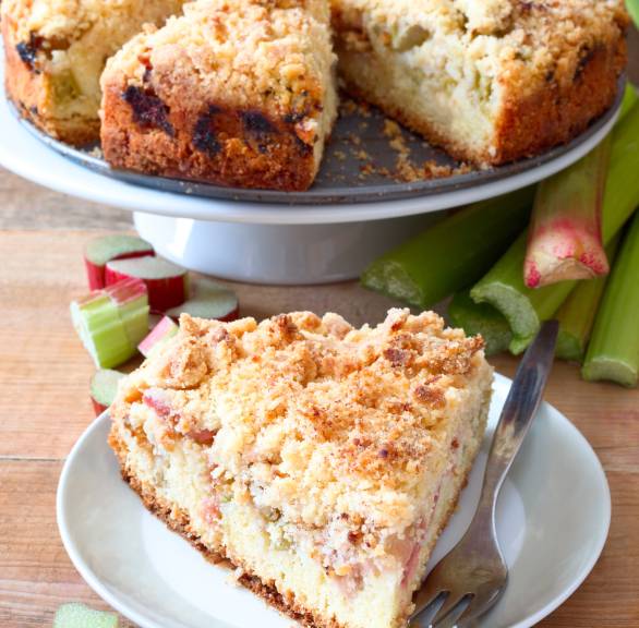 Runder Rhabarber-Streuselkuchen auf einer Kuchenplatte, davor ein Stück auf einem Teller