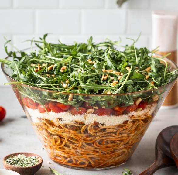 Schichtsalat in einer großen Salatschüssel mit Schichten aus Spaghetti, Mozzarella, Tomaten und Rucola, darauf Pinienkerne