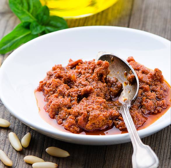 Selbst gemachtes Pesto rosso in einer Schale, daneben Pinienkerne und Olivenöl auf einem Holztisch