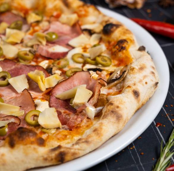 Pizza Capricciosa auf einem Pizzateller belegt mit Mozzarella, Artischocken, Champignons, Schinken und Oliven vor einem Backofen auf einem Tisch