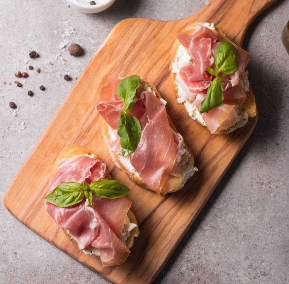 Drei Bruschettas mit Frischkäse und Schinken belegt auf einem Holzbrett garniert mit frischem Basilikum
