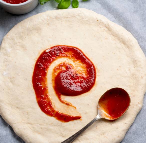 Glutenfreier Pizzateig ausgerollt als runder Pizzaboden wird mit Tomatensauce bestrichen, daneben frischer Basilikum
