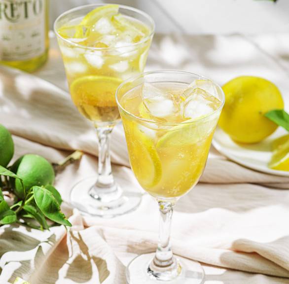 Limoncello Spritz mit Eiswürfeln in zwei Cocktailgläsern, daneben Zitronen, eine Flasche Limoncello und Zitronenmelisse auf einem Tisch