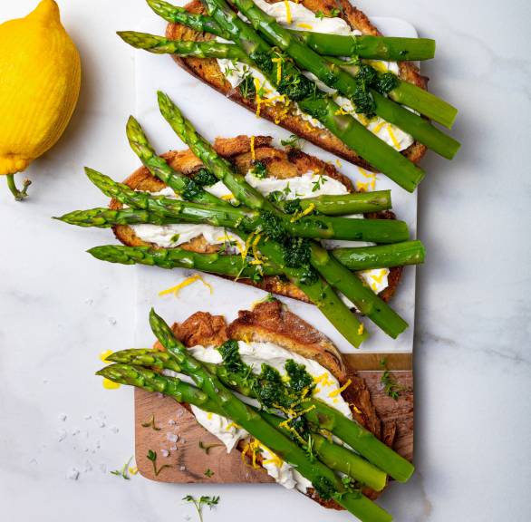 Drei Spargel-Ricotta-Bruschette auf einem Holzbrett getoppt von grünem PEsto und Zitronenabrieb