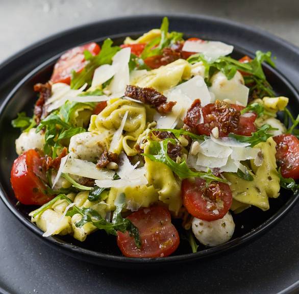 Tortellini-Salat mit Tomaten und Mozzarella auf einem Teller, daneben Rucola und Parmesan