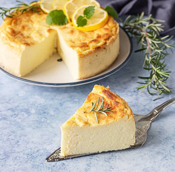 Zitronen-Ricotta-Kuchen (Torta di Ricotta e Limone) angeschnitten auf einer Kuchenplatte garniert mit Zireonenscheiben, Minzblättern und Rosmarin