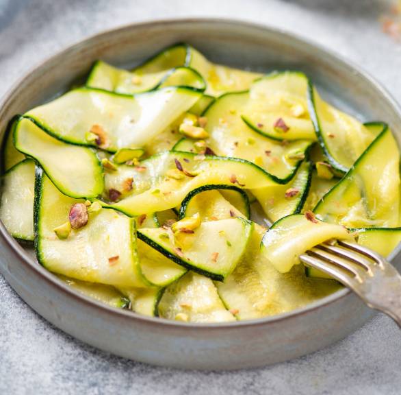 Rohes Zucchini Carpaccio in einer Schale getoppt von Parmesan und Pinienkernen, daneben eine Karaffe mit Olivenöl