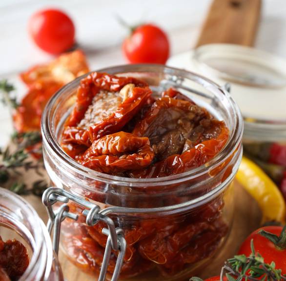 Eingelegte getrocknete Tomaten in Öl in einem Weckglas, daneben Knoblauch, frische Tomaten und Kräuter 