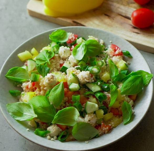 Couscous-Salat mit Paprika, Gurke, Tomate und Mozzarella in einem tiefen Teller, daneben Zutaten auf einem Tisch