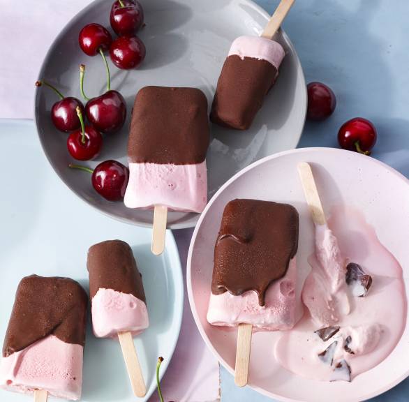 Selbstgemachtes Kirsch-Joghurt-Eis am Stiel mit Schokoglasur verteilt auf mehrere Teller, daneben frische Kirschen