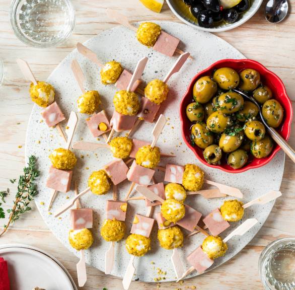 Mortadella-Mozzarella-Spieße auf einem runden Brett, daneben Oliven und Ciabatta