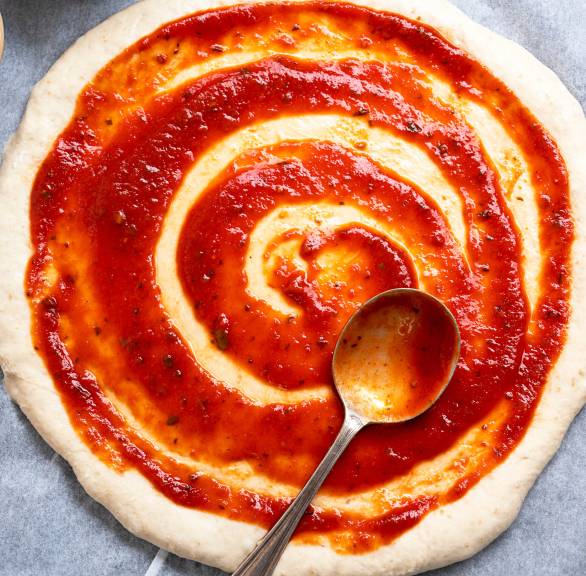 Selbst gemachte Pizzasoße wir mit einem Esslöffel auf Pizzateig gestrichen