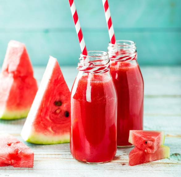 Wassermelonendrink in zwei Glasflaschen mit Strohhalmen, daneben Melonenstücke auf einem Tisch