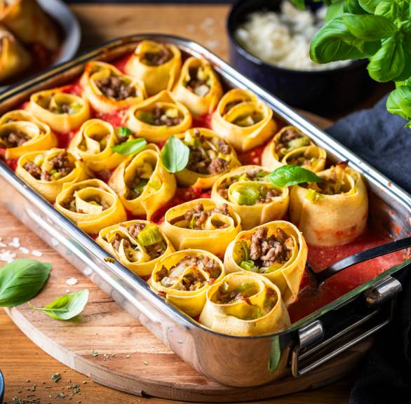 Eine Auflaufform mit gebackenen Pastaschnecken gefüllt mit Lauch und Hackfleisch, daneben frischer Basilikum
