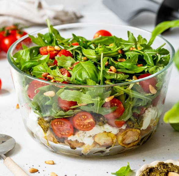 Italienischer Katoffelsalat mit Tomaten, Mozzarella und Rucola geschichtet in einer Glasschüssel, daneben grünes Pesto auf einem Tisch