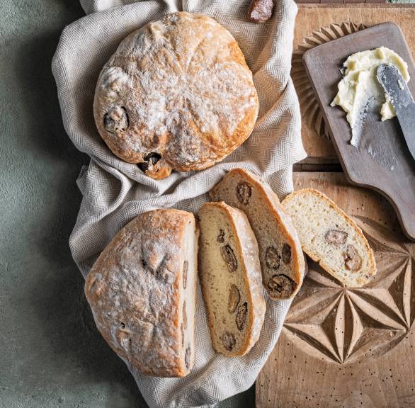 Zwei Laibe Maronenenbrot auf einem Tisch, eines angeschnitten, daneben gemusterte Holzbretter