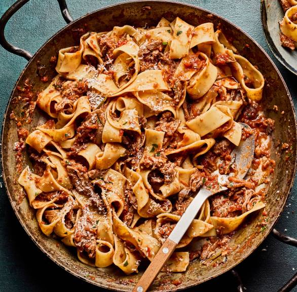 Pappardelle mit Enten-Ragù in einer tiefen Pfanne auf einem festlich dekorierten Tisch, daneben Parmesan