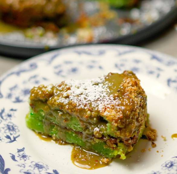 Ein Stück Pistazienkuchen auf einem Teller, dahinter der restliche Kuchen auf einer Kuchenplatte