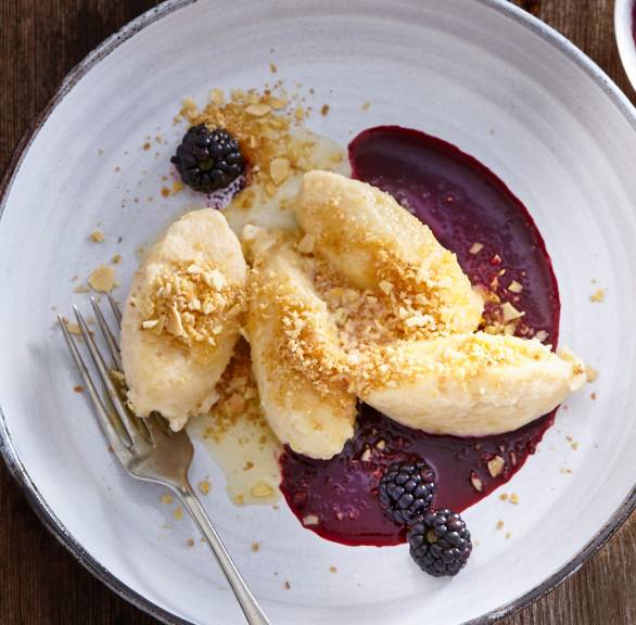 Vier Hüttenkäse-Grießnocken auf einem Dessertteller mit Brombeersauce bestreut mit Mandel-Bröseln