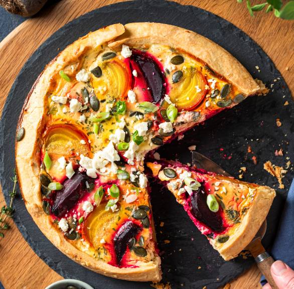 Runde Quiche mit roter und gelber Bete, Feta und Kürbiskernen angeschnitten auf einer Schieferplatte von oben