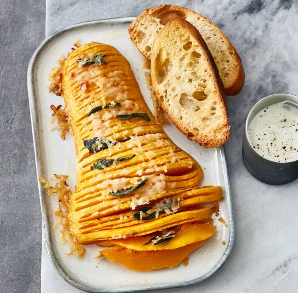 Halbierter, eingeschnittener Butternut-Kürbis überbacken mit Käse auf einem Teller, daneben Ciabattascheiben und Dip