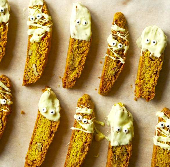 Mehrere Kürbis-Cantuccini nebeneinander auf einem Backblech dekoriert mit weißer Schokolade und Zuckeruagen als Mumien-Kekse