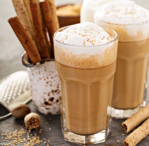 Zwei hohe Gläser mit Latte Macchiato auf einem Tisch, daneben Zimtstangen und Gewürze auf einem Tisch
