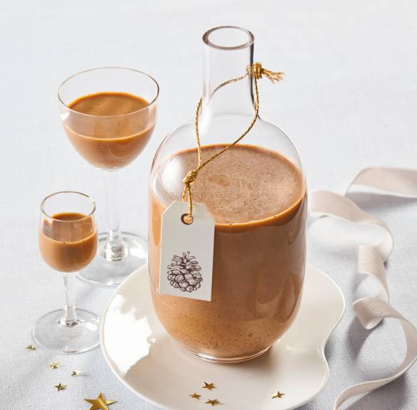 Selbstgemachter Zimtlikör in einer Glasflasche, daneben zwei volle Schnapsgläser auf einem weihnachtlich dekorierten Tisch