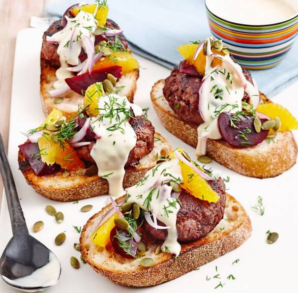Mehrere geröstete Ciabattascheiben belegt mit Polpette, Roter Bete, Orangenfilets getoppt mit Joghurtsoße und Dill auf einer Servierplatte