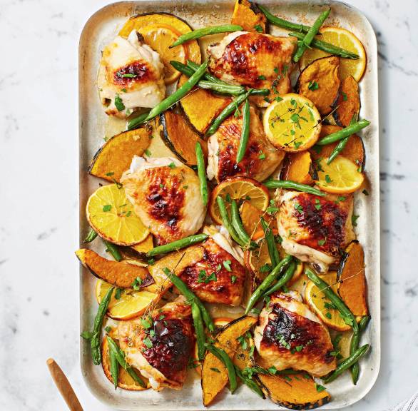Backblech mit Hähnchenkeulen, Kürbisspalten, grünen Bohnen und Orangenscheiben auf einem Tisch