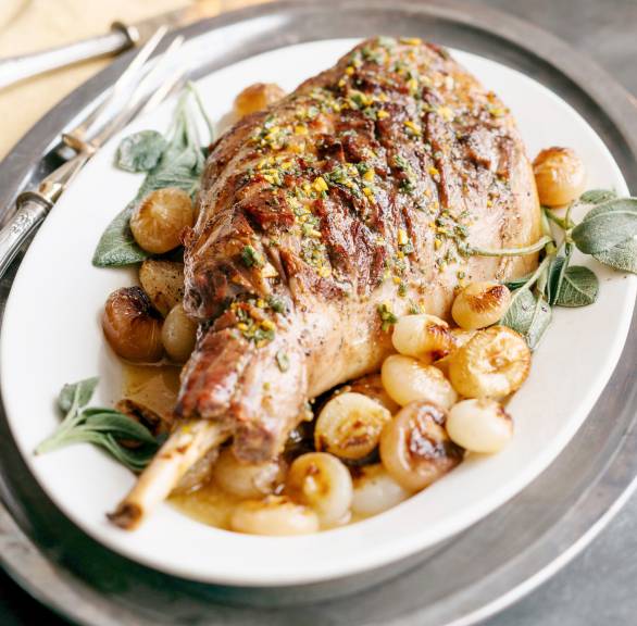 Eine gebratene Lammkeule bestrichen mit mit Senf-Gremolata auf Perlzwiebelgemüseangerichtet auf einem Teller