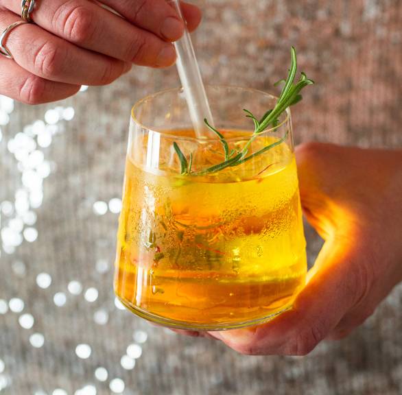 Goldener Gin Tonic mit Safran garniert mit einem Rosmarinzwei in einem Gin Tonic Glas in der Hand einer Frau mit einem Pailettenoberteil