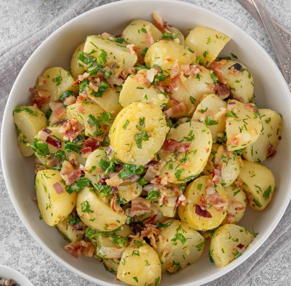 Kartoffelsalat mit gebratenen Pancetta-Streifen in einer Salatschüssel auf einem Tisch, daneben Olivenöl und frische Kräuter