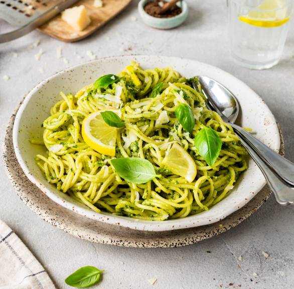 Ein Teller Spaghetti mit grünem Pesto und Zitronen auf einem Tisch, daneben Parmesan