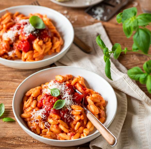 Nudeln mit Serena Loddos Tomatensoße auf zwei Pastatellern, daneben Käse und Tomaten auf einem Holztisch
