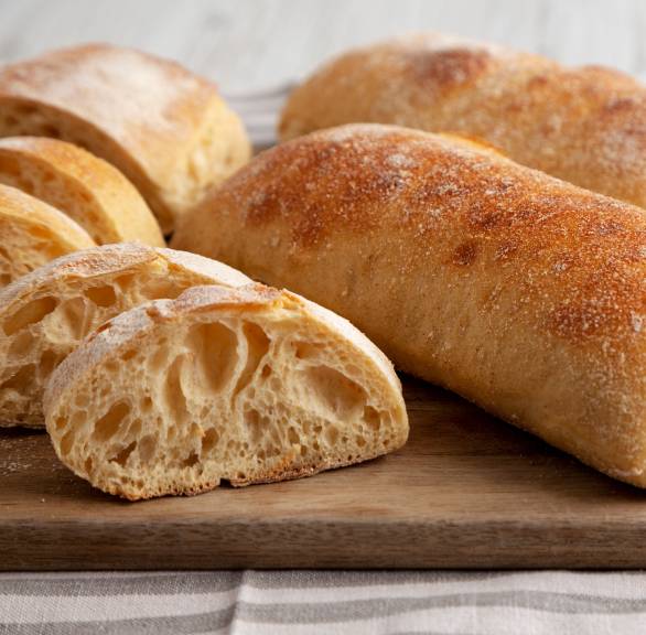 Drei selbstgebackene Ciabatta-Brote mit Trockenhefe auf einem Holzbrett, eines davon in Scheiben geschnitten