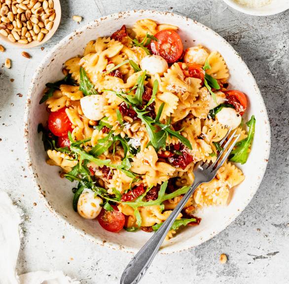 Nudelsalat mit getrockneten Tomaten und Mozzarella angerichtet mit Rucola und Pinienkernen auf einem tiefen Teller