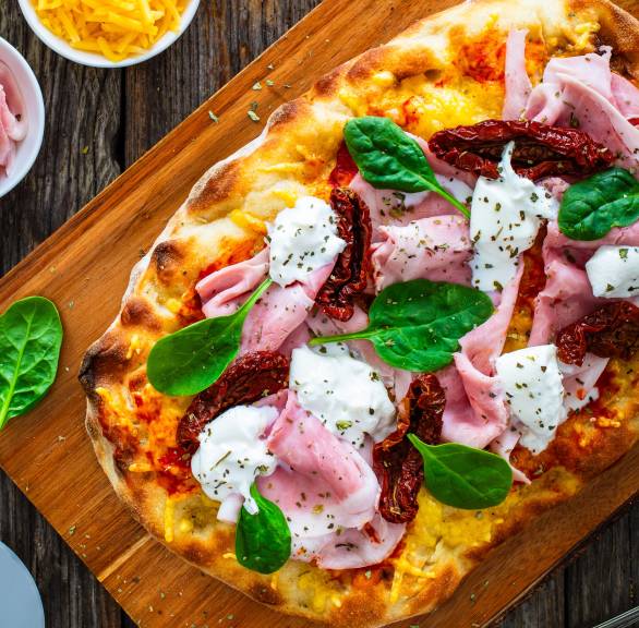 Gebackene, ovale Pinsa belegt mit getrockneten Tomaten, gekochtem Schinken und Burrata auf einem Holzbrett