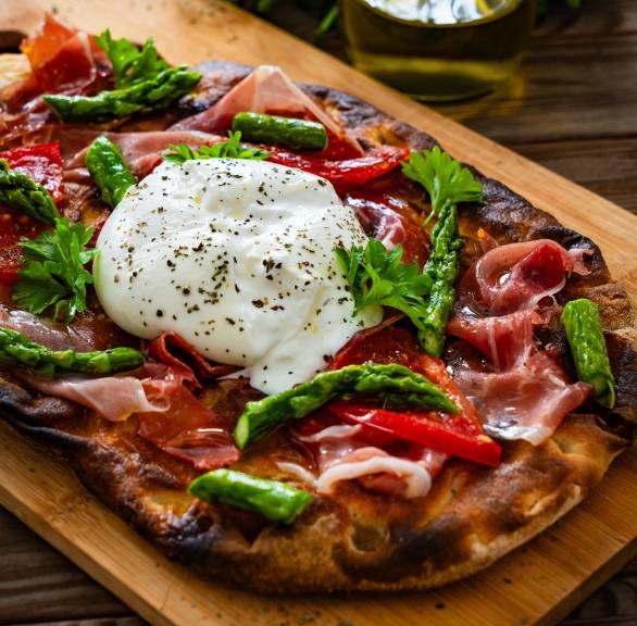 Selbstgebackene Pinsa belegt mit grünem Spargel, Schinken und Burrata auf einem Holzbrett