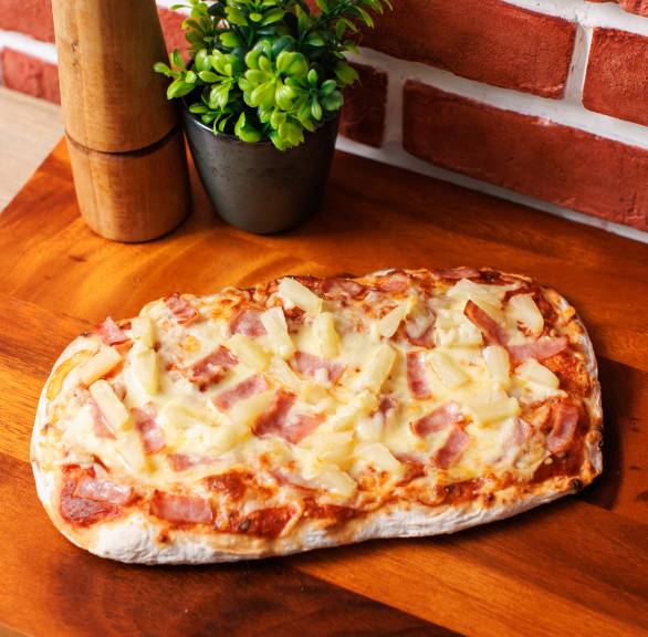 Eine gebackene Pinsa Hawaii mit Mozzarella, Schinken und Ananas auf einem Holzbrett