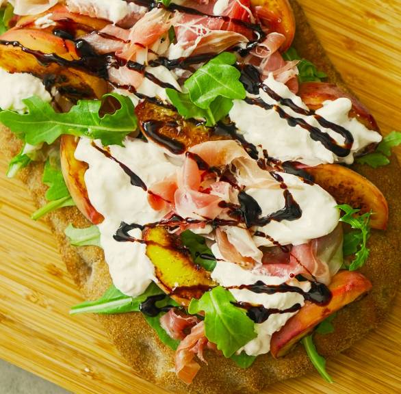 Gebackene Pinsa belegt mit gegrilltem Pfirsich, Burrata und Parmaschinken auf einem Holzbrett