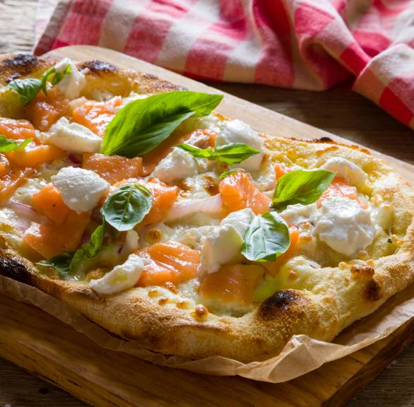 Ganze gebackene Pinsa Bianca mit Lachs und Ricotta getoppt von frischen Basilikumblättern auf einem Holzbrett