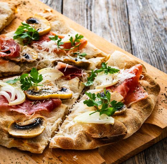 Pinsa Bianca belegt mit Champignons, Mozzarella und Parmaschinken in Streifen geschnitten auf einem Holzbrett
