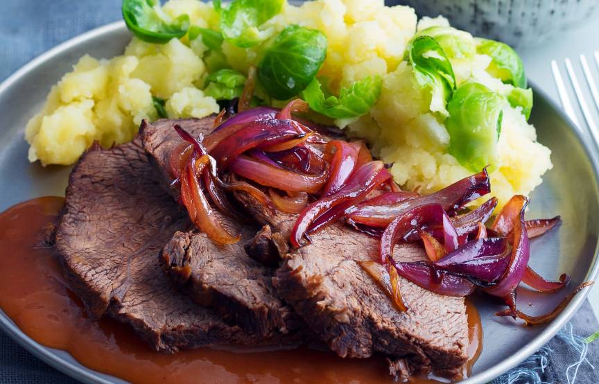 Scheiben von Rinderbraten auf einem Teller mit Sauce bedeckt, daneben Kartoffelpüree und Rosenkohl