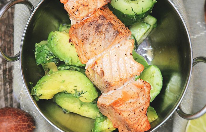 Gegrillter Lachs auf einem Spieß auf Gurkensalat, daneben Avocadodip und halbierte Avocados